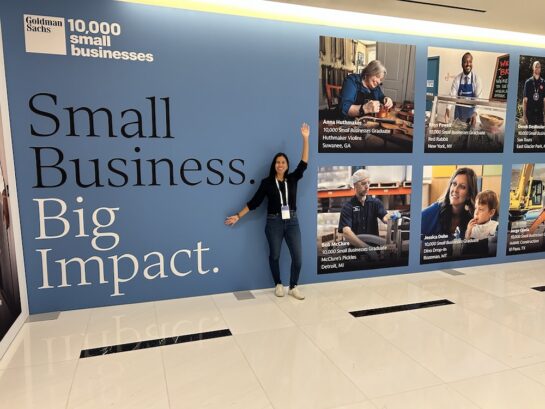 A woman stands in front of a large blue wall display with the text "Small Business. Big Impact." and the Goldman Sachs 10,000 Small Businesses logo. The wall also features photos and names of small business graduates with their business names and locations, including Anna Huthmaker, Rhys Powell, Bob McClure, Jessica Dehn, and Jorge Ojeda. The woman is smiling, wearing a black top, blue jeans, white sneakers, and a lanyard with badges, raising one arm. The floor is white with black accents.